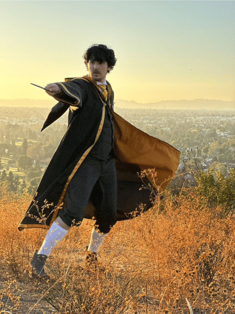 Person dressed as wizard, pointing wand on top of grassy hill, overlooking valley at sunset