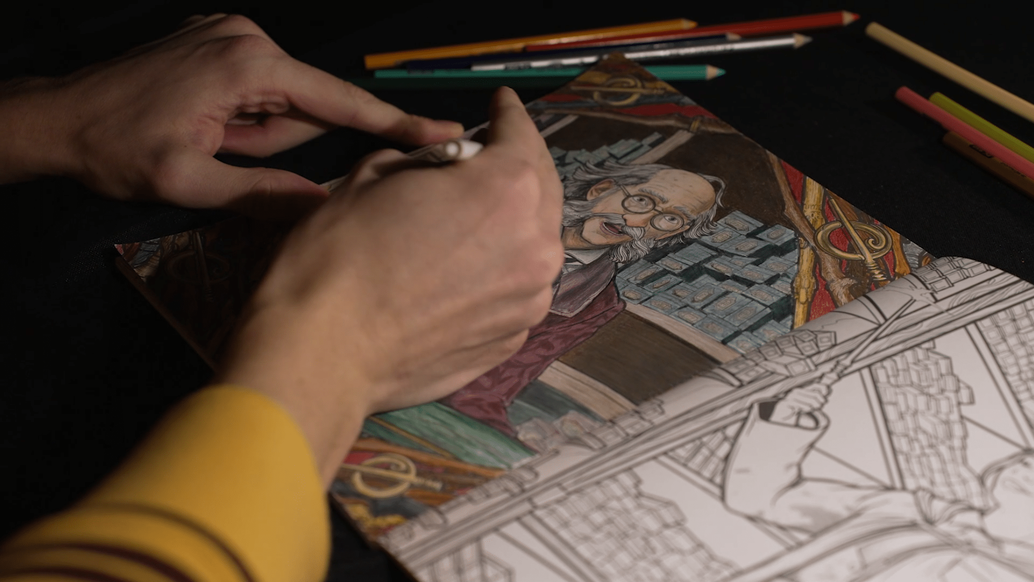 The image shows a close-up of hands coloring a page in a detailed coloring book under soft, focused lighting.Left Page: The page is partially visible, showing the line art of an illustrated scene that is not yet colored. The artwork includes a figure holding a wand or staff and architectural elements, hinting at a magical or fantasy theme.Right Page: The page is almost completely colored and depicts an older man with a bushy mustache and round glasses, wearing a burgundy robe or jacket. The background shows stacks of books or boxes and some intricate decorative elements, such as golden rings or magical tools, indicating a study or library setting. The coloring is carefully done with various shades of brown, red, green, and blue, giving depth and texture to the scene.A hogwarts legacy artist wearing a yellow and brown striped shirt is actively coloring the page with a white or light-colored pencil. Other colored pencils are scattered above the book on the black tabletop, suggesting ongoing work and creativity.